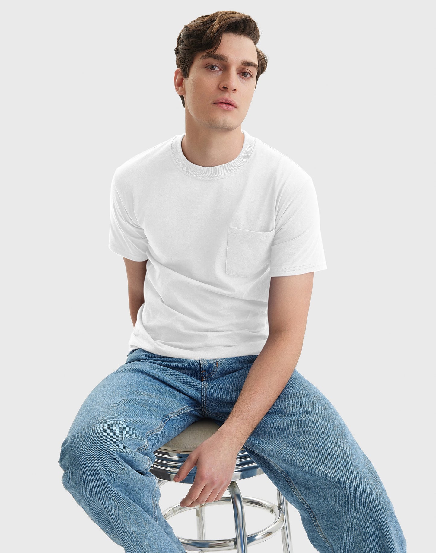 Man sitting on a stool wearing a white t-shirt and blue jeans against a plain background