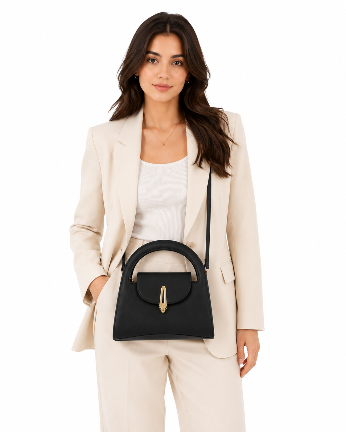 A woman wearing a cream-colored blazer and matching trousers models a black structured handbag with a top handle and gold-tone hardware against a plain white background.