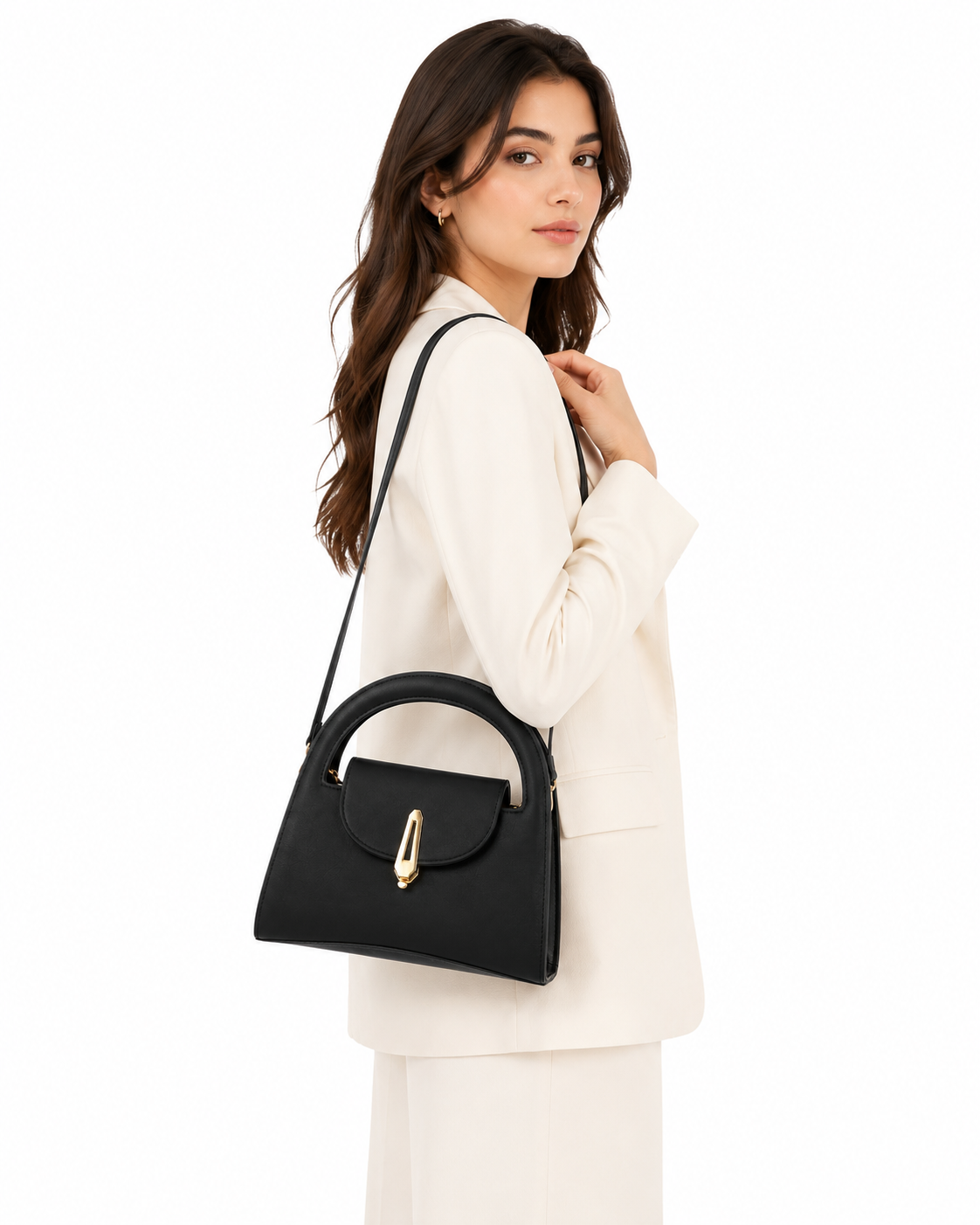 A woman in a cream-colored blazer and matching trousers models a black leather shoulder bag with a gold-tone turn-lock clasp and top handle against a plain white background.