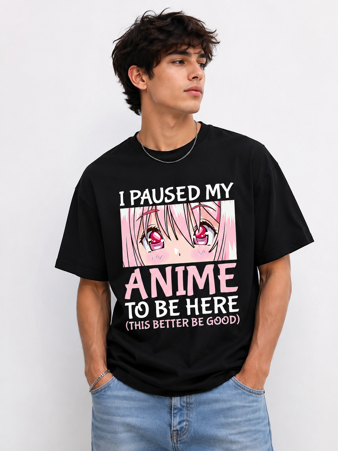 A man wearing a black graphic t-shirt with anime eyes and the text 'I PAUSED MY ANIME TO BE HERE (THIS BETTER BE GOOD)' paired with light blue jeans, against a plain light gray background.