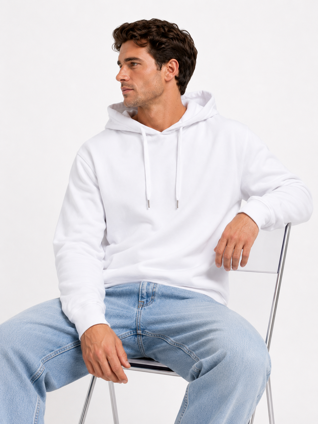 A man wearing a plain white pullover hoodie and light blue jeans, seated on a minimalist metal chair against a clean white studio background.