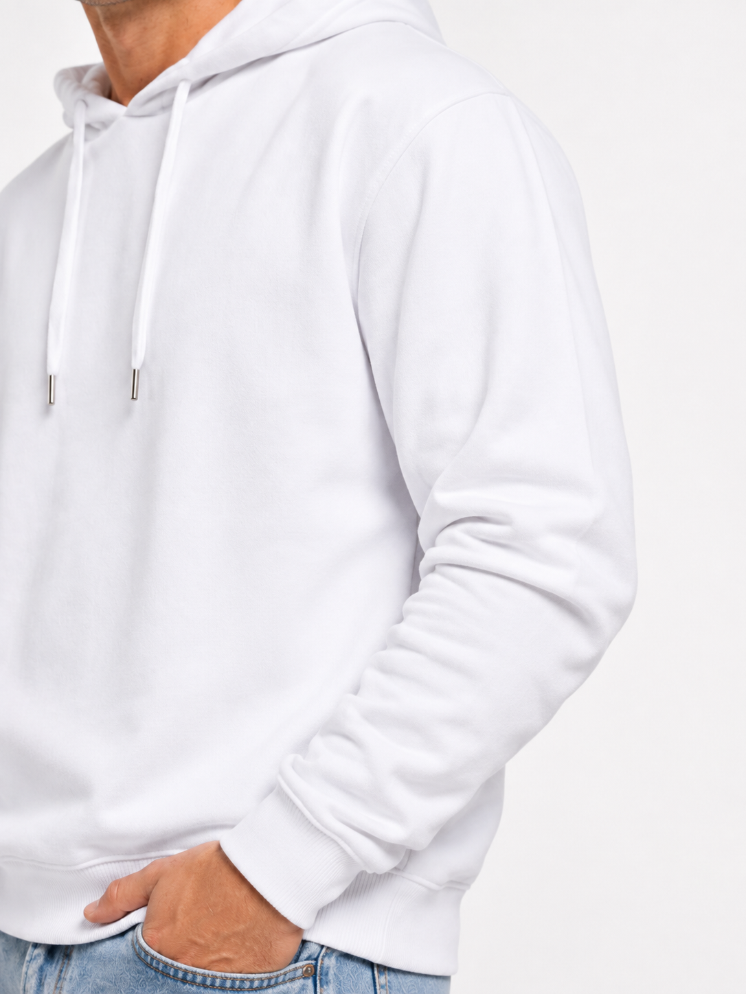 A close-up of a person wearing a plain white pullover hoodie with white drawstrings and ribbed cuffs, paired with light blue jeans against a clean white background.