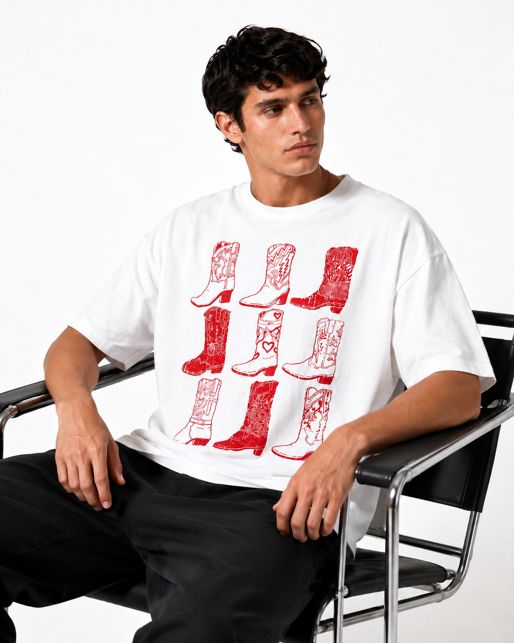 A man wearing a white oversized t-shirt with a red graphic print of cowboy boots, seated on a black metal chair against a white studio background.