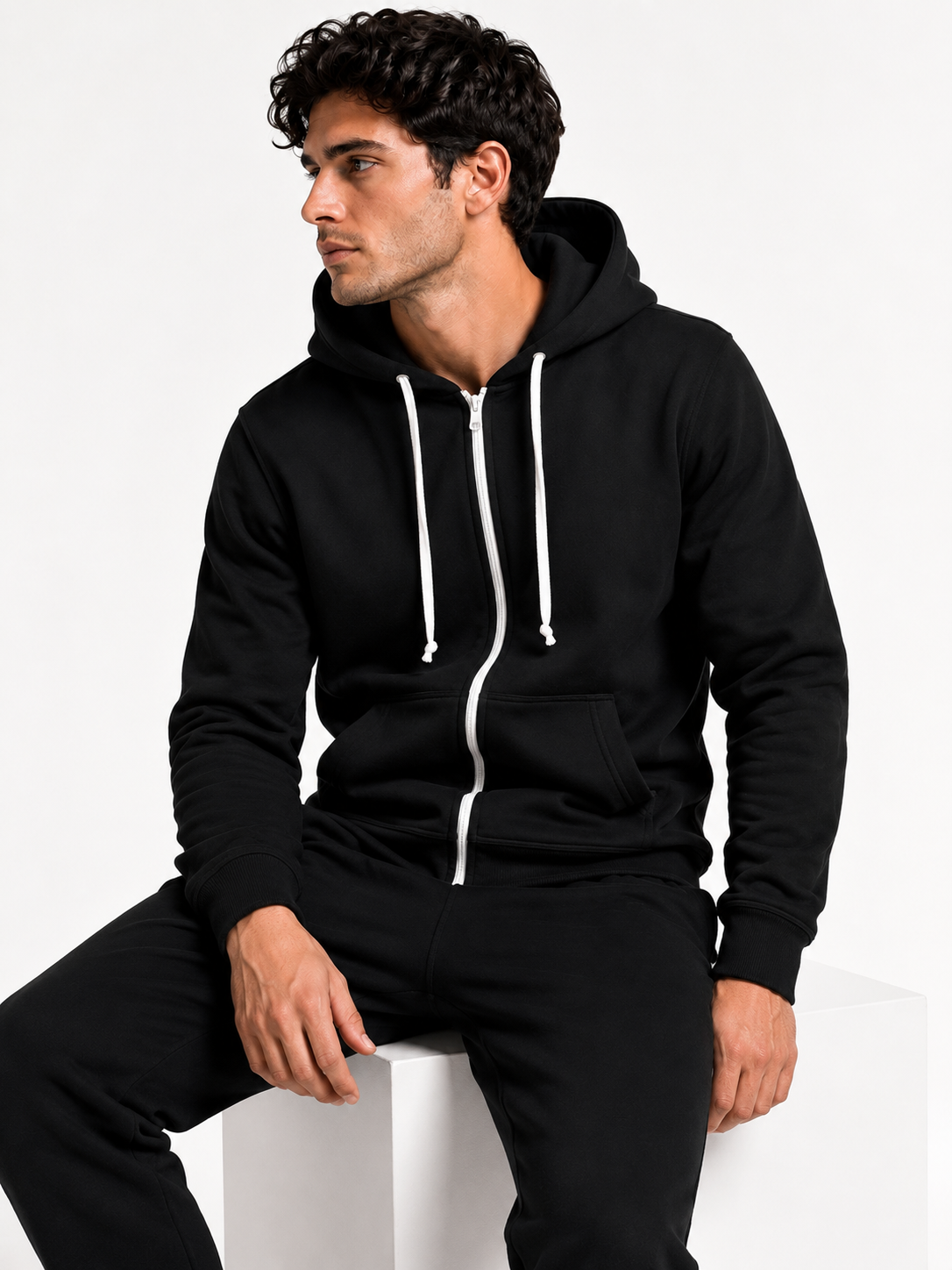 A man wearing a black zip-up hoodie with white drawstrings and white zipper accents, paired with black sweatpants, sitting on a white block against a plain white background.